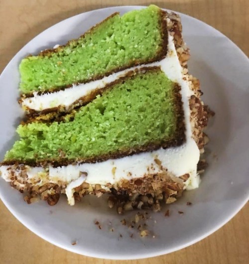 A slice of green cake with white frosting and a nutty crust, served on a white plate.