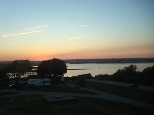 Sunset over a calm lake, with silhouettes of trees and a few vehicles parked nearby. Soft colors in the sky.