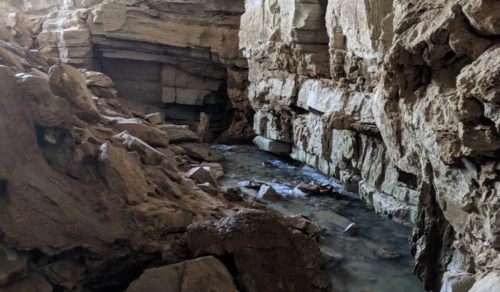A rocky cave interior with a small stream flowing through, surrounded by large stone formations.