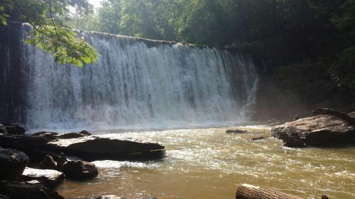A serene waterfall cascades over rocks into a calm river, surrounded by lush green trees and mist.