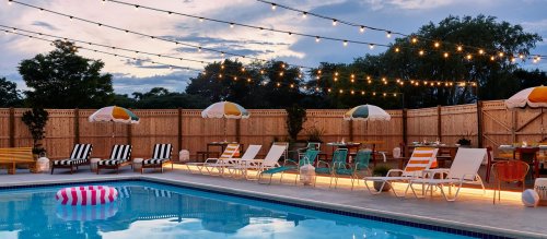 A serene poolside scene with lounge chairs, colorful umbrellas, and string lights illuminating the evening sky.