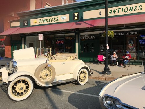 A vintage white convertible parked outside a quaint antique shop, with people sitting nearby enjoying the day.