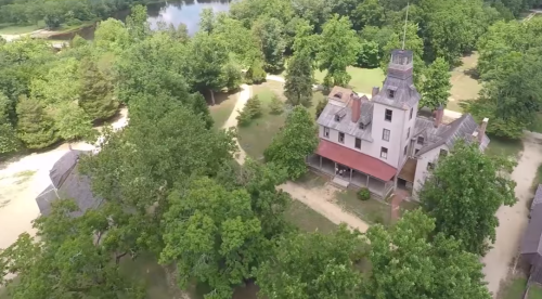 Aerial view of a large, historic house surrounded by lush greenery and pathways, near a calm body of water.
