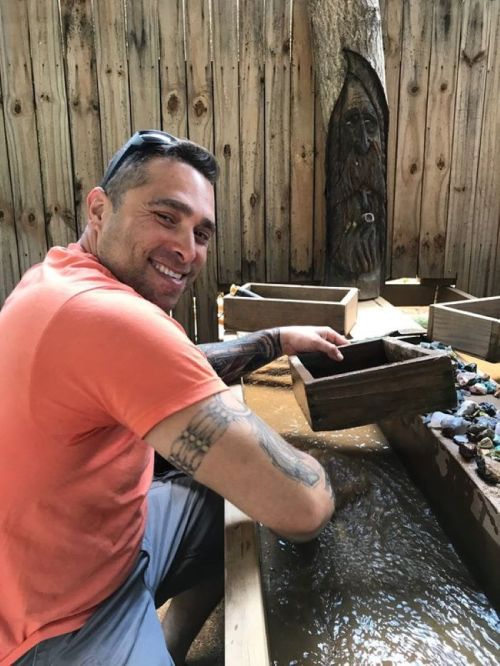 A smiling man in an orange shirt sifts through a wooden box in a water-filled area, surrounded by wooden structures.