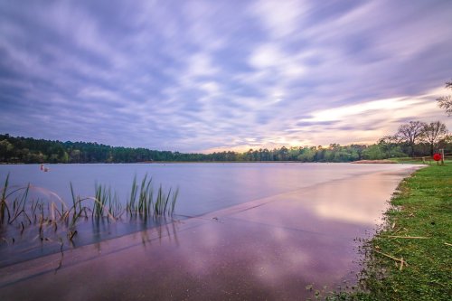 A serene lake at dusk, with smooth water reflecting the sky and lush greenery along the shore.