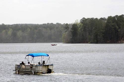 A pontoon boat with people on a lake, with trees lining the shore and another boat in the distance.