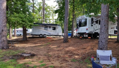 Two RVs parked among tall trees at a campsite, with a fire pit and a camping chair in the foreground.