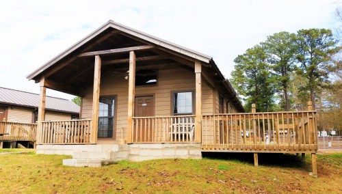 A wooden cabin with a porch, surrounded by trees, set on a grassy area under a cloudy sky.