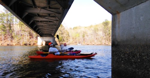 Two kayakers paddle under a bridge on a calm river, surrounded by trees and clear blue skies.