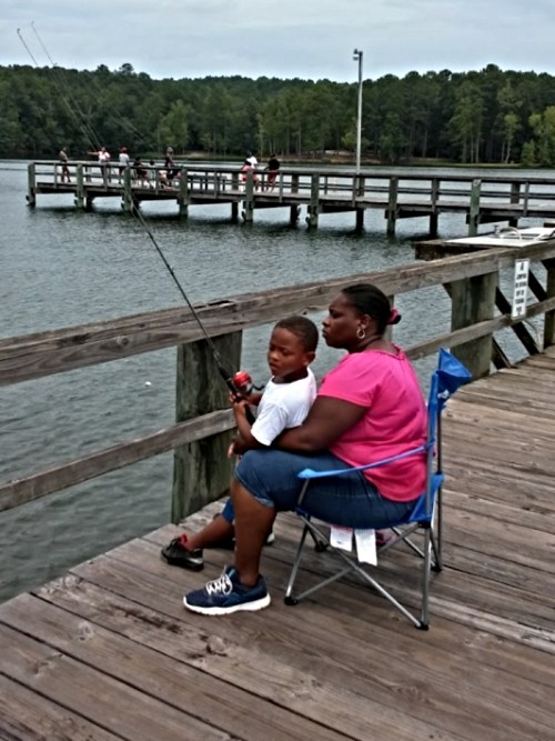 A woman and a boy sit on a dock, fishing together, with a calm lake and trees in the background.