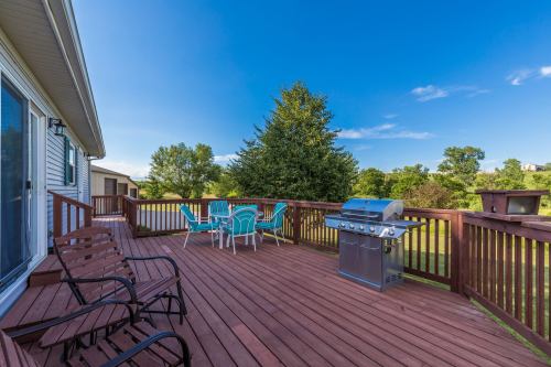 A wooden deck with chairs and a table, featuring a grill, surrounded by greenery and a clear blue sky.
