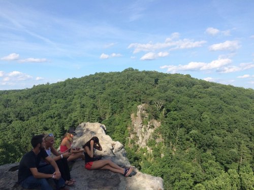A group of people sitting on a rocky ledge, overlooking a lush green hillside under a blue sky with scattered clouds.