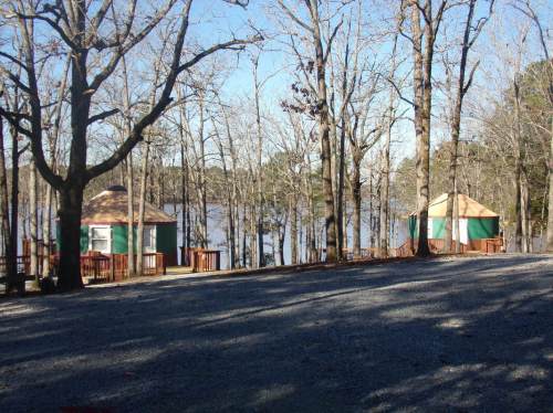 Two round cabins with green and brown exteriors sit by a lake, surrounded by bare trees and a gravel path.