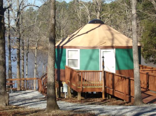A colorful yurt with a wooden deck, surrounded by trees near a calm lake on a sunny day.
