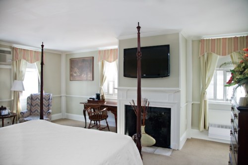 A cozy hotel room featuring a four-poster bed, desk, TV, and large windows with soft curtains.