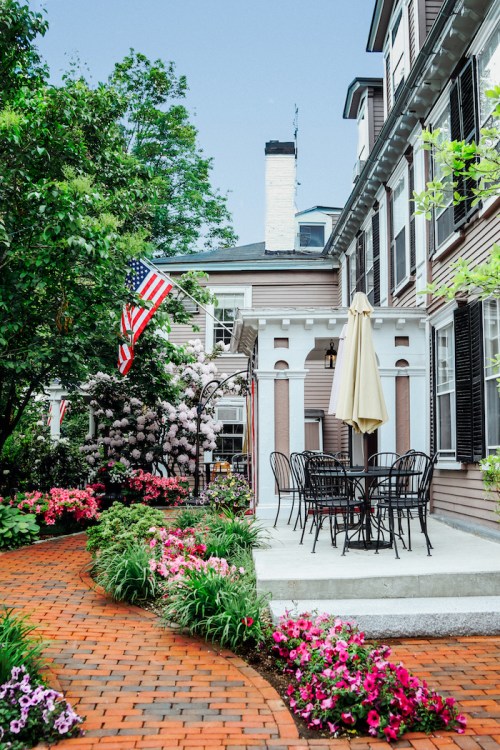 A charming patio with a table and chairs, surrounded by colorful flowers and an American flag, set against a historic building.