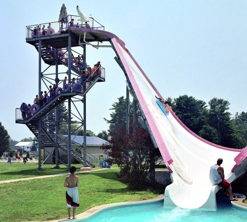 A tall water slide with people waiting at the top and one person sliding down into a pool below.