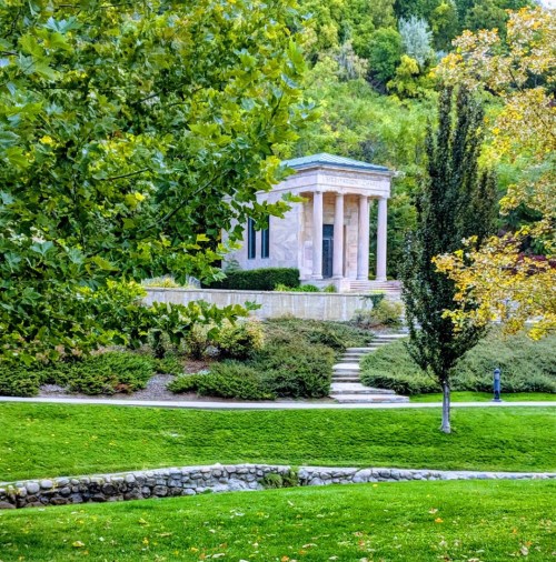A classical stone structure with columns surrounded by greenery and trees in a serene park setting.