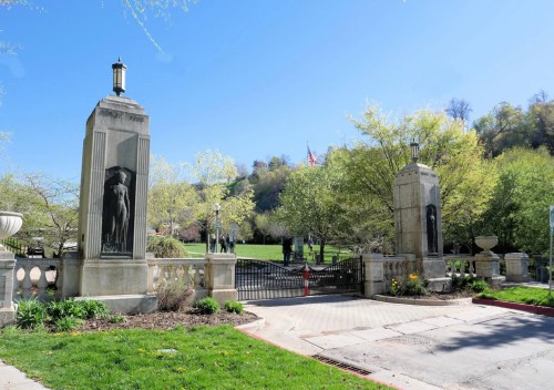 Entrance gate with stone pillars and sculptures, surrounded by greenery and trees under a clear blue sky.