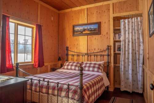Cozy bedroom with wooden walls, a plaid bedspread, red curtains, and a mountain landscape painting.
