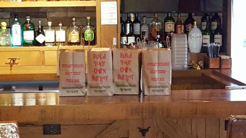 Four takeout bags on a bar counter, each labeled "Build Your Own Burger," with bottles of liquor in the background.