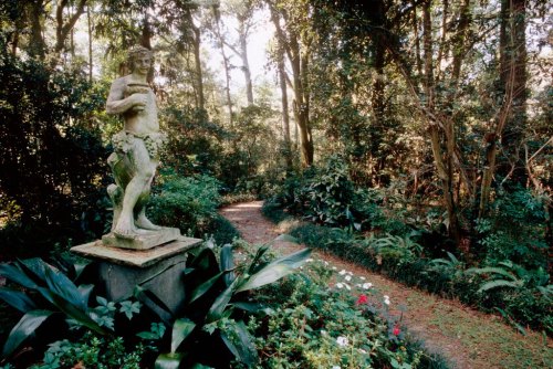 A stone statue stands amidst lush greenery along a winding garden path, surrounded by vibrant flowers and trees.