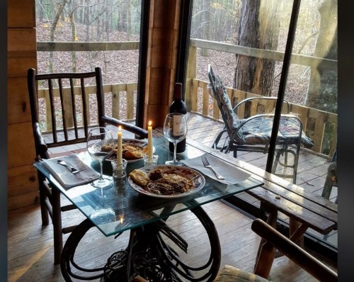 A cozy dining setup with a glass table, plates of food, wine glasses, and candles, overlooking a forest view.