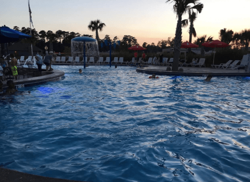 A vibrant pool scene at sunset, with people swimming, lounge chairs, and palm trees in the background.