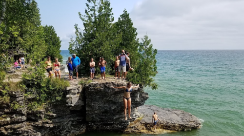 A group of people on a rocky shore by the water, with one person preparing to jump off a ledge into the lake.