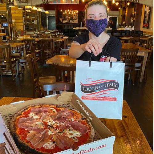 A masked woman holds a takeout bag in a restaurant, with a pizza topped with prosciutto in the foreground.