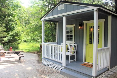 A cozy gray cabin with a yellow door and porch, surrounded by greenery and a picnic area nearby.