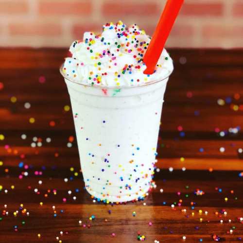 A creamy milkshake topped with whipped cream and colorful sprinkles, served in a clear cup with a red straw.