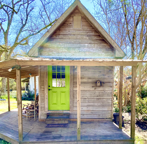 A small wooden cabin with a bright green door and porch, surrounded by trees and greenery.
