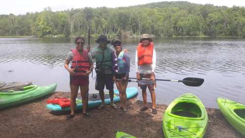 Four people in life jackets stand by a calm lake with kayaks, surrounded by lush greenery.