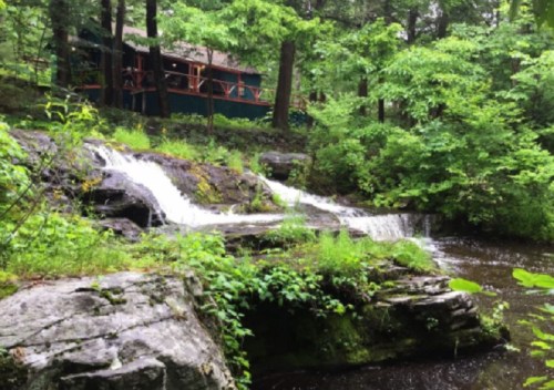 A serene scene of a small waterfall flowing over rocks, surrounded by lush greenery and a cabin in the background.