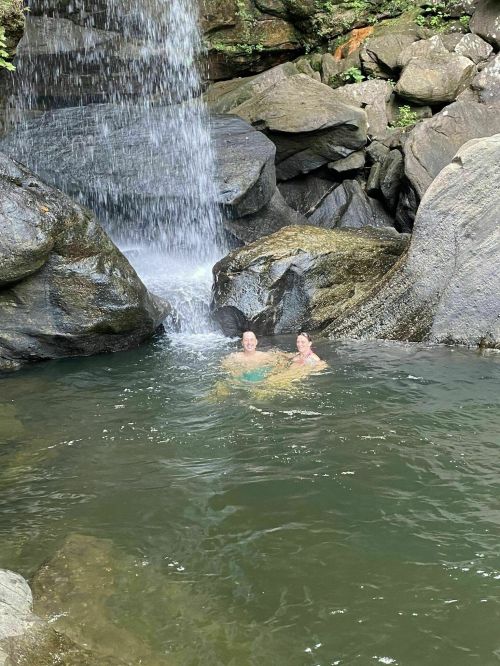 Two people relax in a natural pool beneath a waterfall, surrounded by rocks and lush greenery.