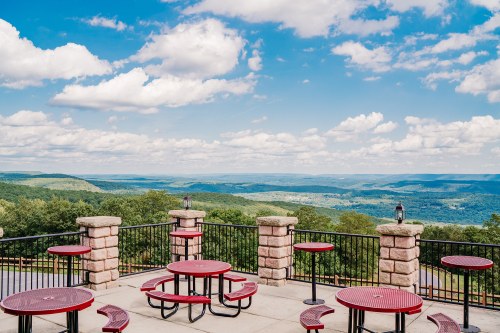 A scenic view from a terrace with red picnic tables overlooking green hills and a blue sky with fluffy clouds.