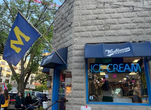 A corner ice cream shop with a blue flag and neon "ICE CREAM" sign, surrounded by trees and people enjoying the area.