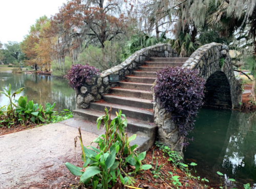 A stone bridge with purple plants spans a tranquil pond, surrounded by lush greenery and trees.