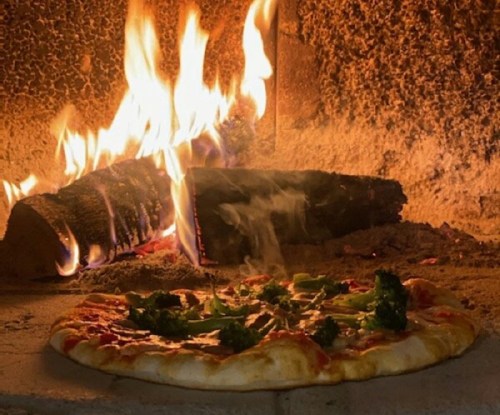 A freshly baked pizza with toppings sits in front of a roaring wood-fired oven.