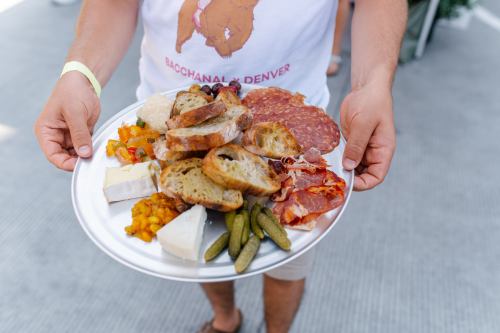 A person holds a plate filled with assorted cheeses, meats, bread, pickles, and colorful vegetables.