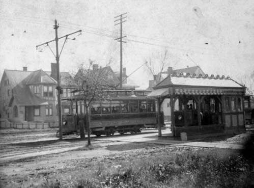 Historic black-and-white photo of a streetcar at a station, with houses and trees in the background.