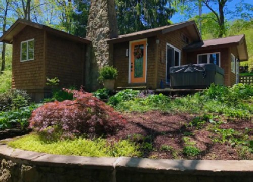 A cozy cabin surrounded by lush greenery and a stone wall, featuring a hot tub and colorful plants in the foreground.