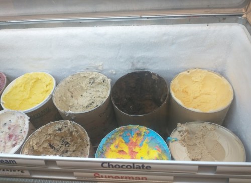 A variety of ice cream flavors in metal containers, including yellow, brown, and colorful options, displayed in a freezer.