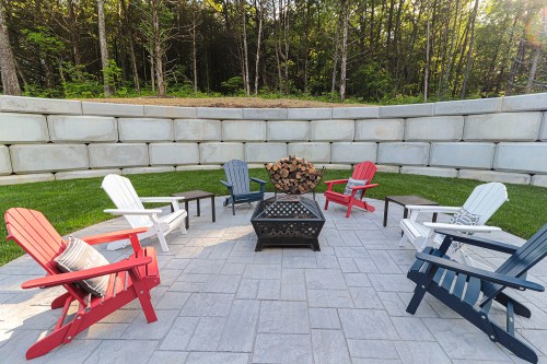 A circular patio with colorful Adirondack chairs around a fire pit, surrounded by trees and stone walls.