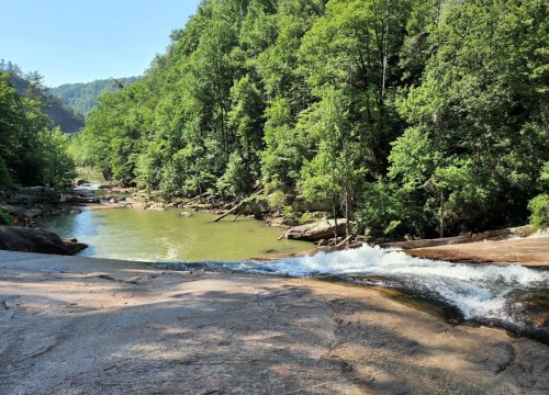 A serene river flows over smooth rocks, surrounded by lush green trees and hills under a clear blue sky.