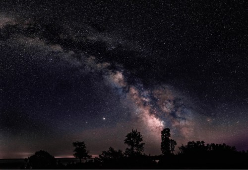 A stunning view of the Milky Way galaxy over a dark landscape, with silhouettes of trees against a starry sky.