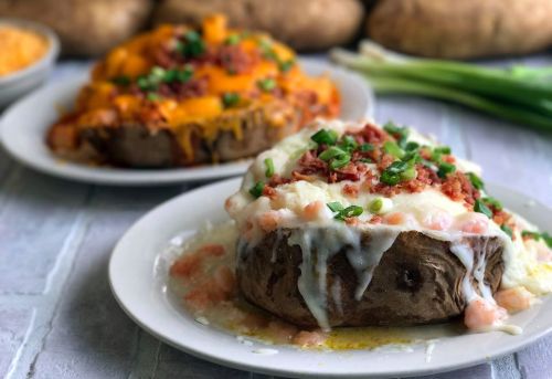Two loaded baked potatoes on white plates, one topped with cheese and bacon, the other with shrimp and green onions.