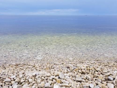 A calm, clear sea meets a rocky shoreline under a cloudy sky.