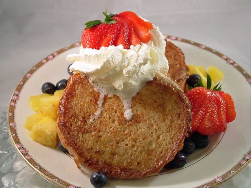 A plate of French toast topped with whipped cream and strawberries, surrounded by blueberries and pineapple chunks.
